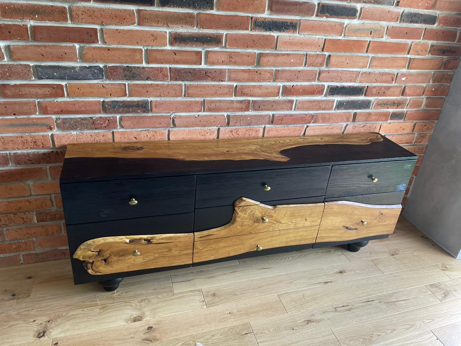 Prezentacja designerskiej komody z litego drewna z zachowaną naturalną krawędzią oraz czarnym wykończeniem, jako przykładu nowoczesnego wzornictwa w aranżacji wnętrz.