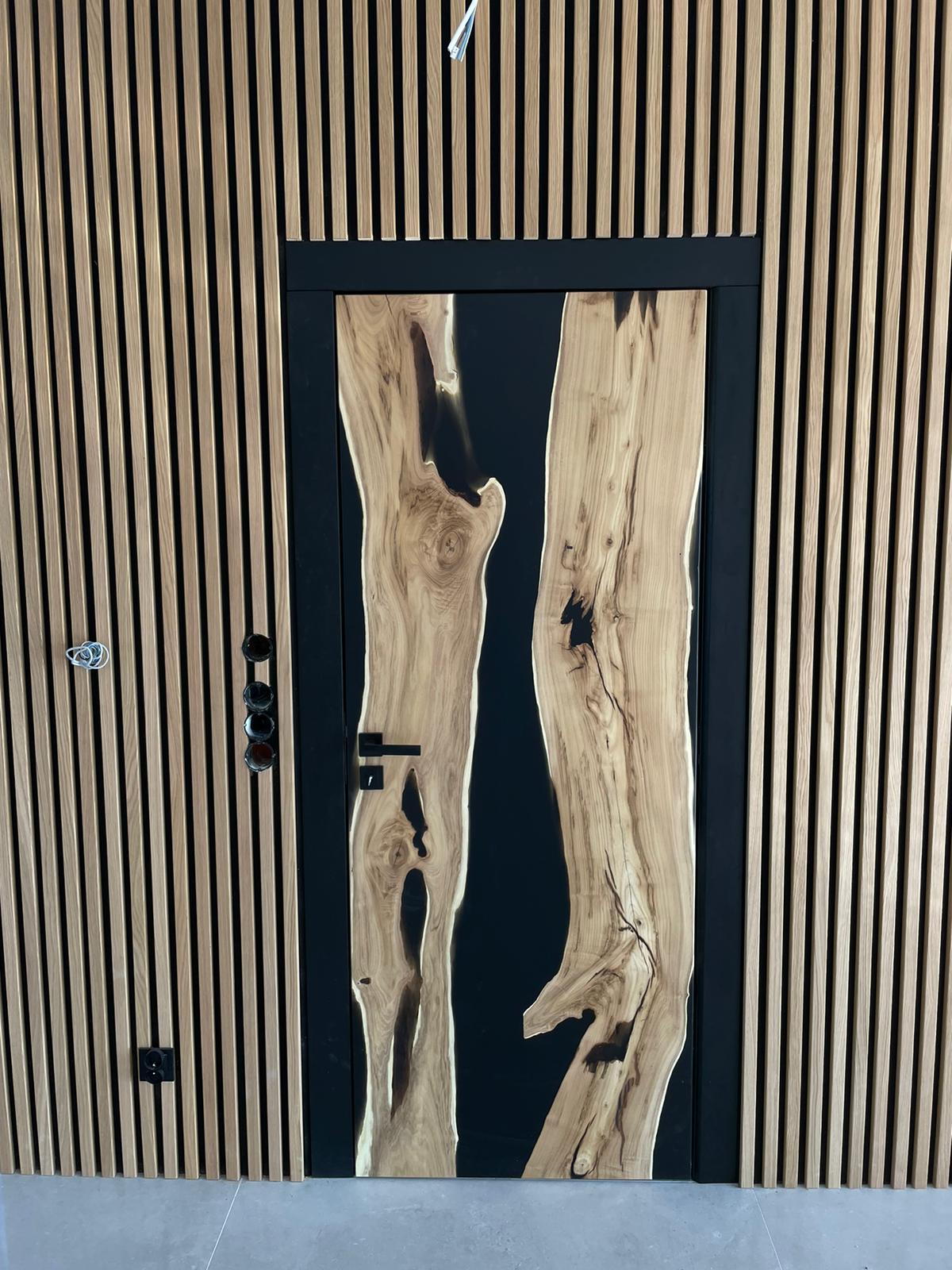 Prezentacja nowoczesnych drzwi wewnętrznych z litego drewna i czarnej żywicy epoksydowej, wkomponowanych w ścianę wykończoną pionowymi lamelami, jako spójny element aranżacji wnętrza.